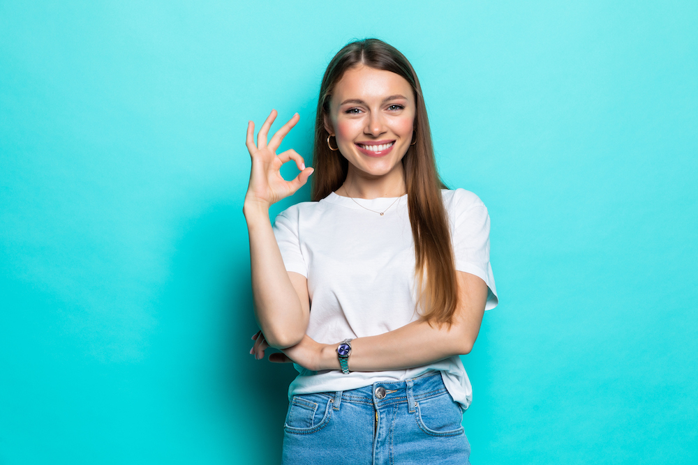 Asian happy portrait beautiful young woman standing smiling showing hand okay sign and looking to camera isolated on pastel blue background with copy space for text