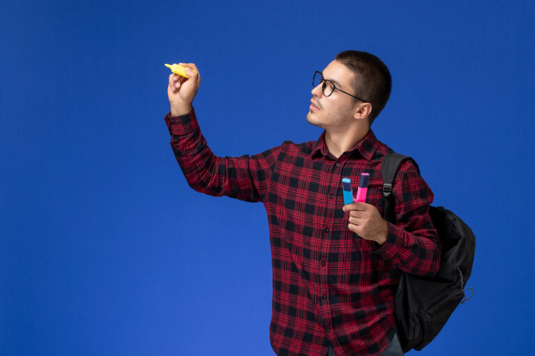 front view male student in red checkered shirt with backpack holding felt pens on light blue background college university lesson male